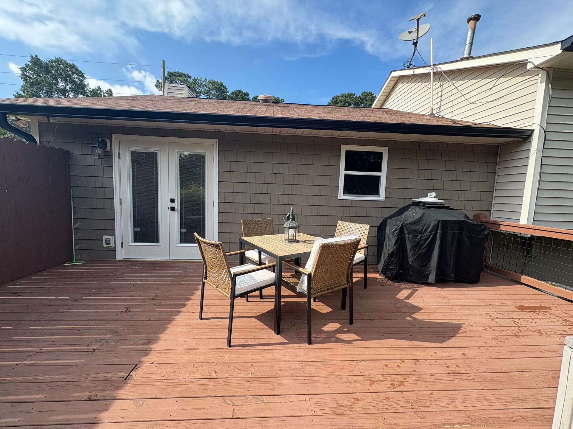 Wooden deck with outdoor furniture, and a grill, against a house with a door, window, and brown siding.