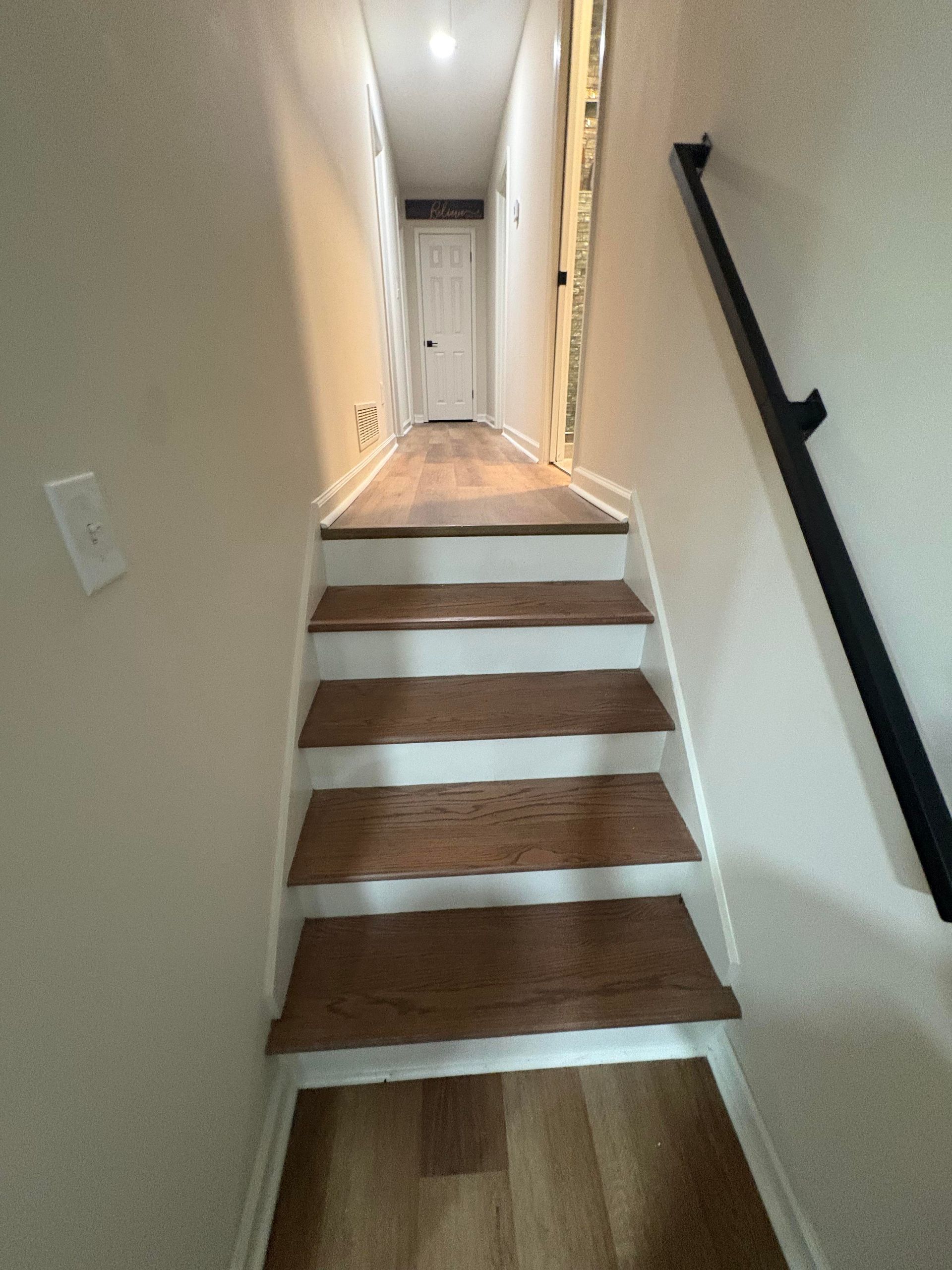 Staircase with wooden steps and white risers, leading to a hallway with a closed door. Black handrail on right.