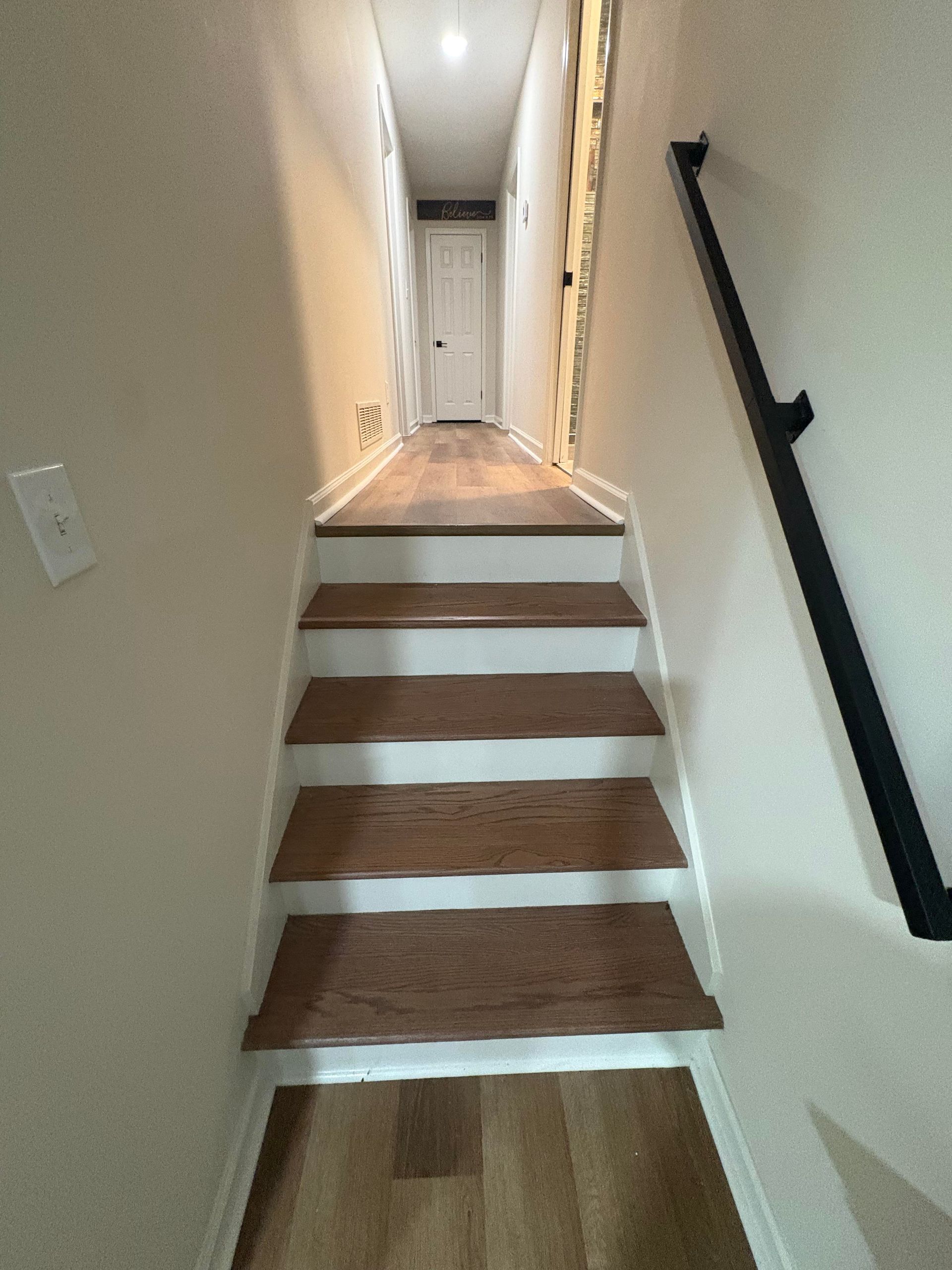 Staircase with wood steps and white risers leading to a hallway with a closed door, black handrail on the right.