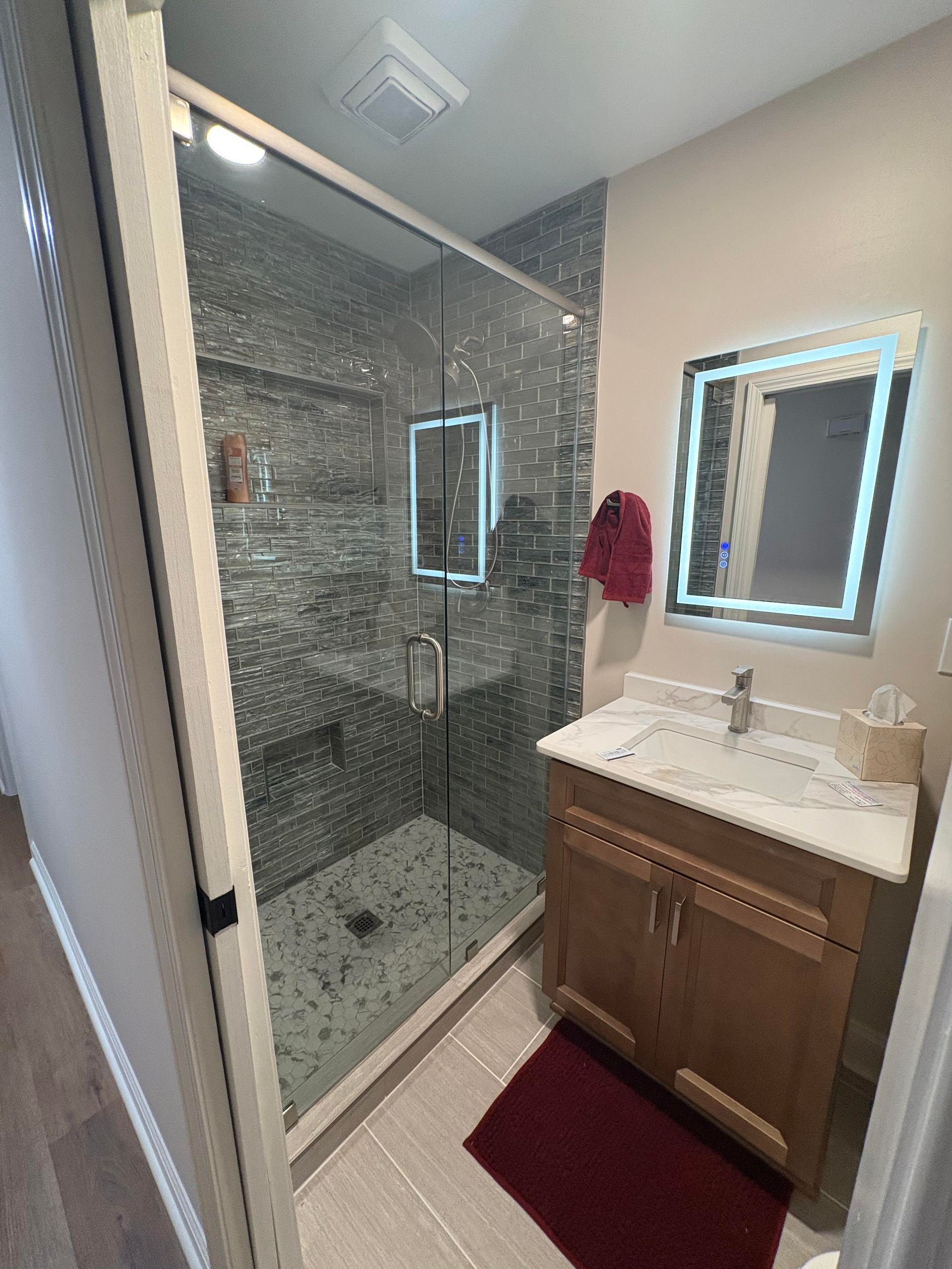 Bathroom with glass shower, wooden vanity, lighted mirror, and tile.