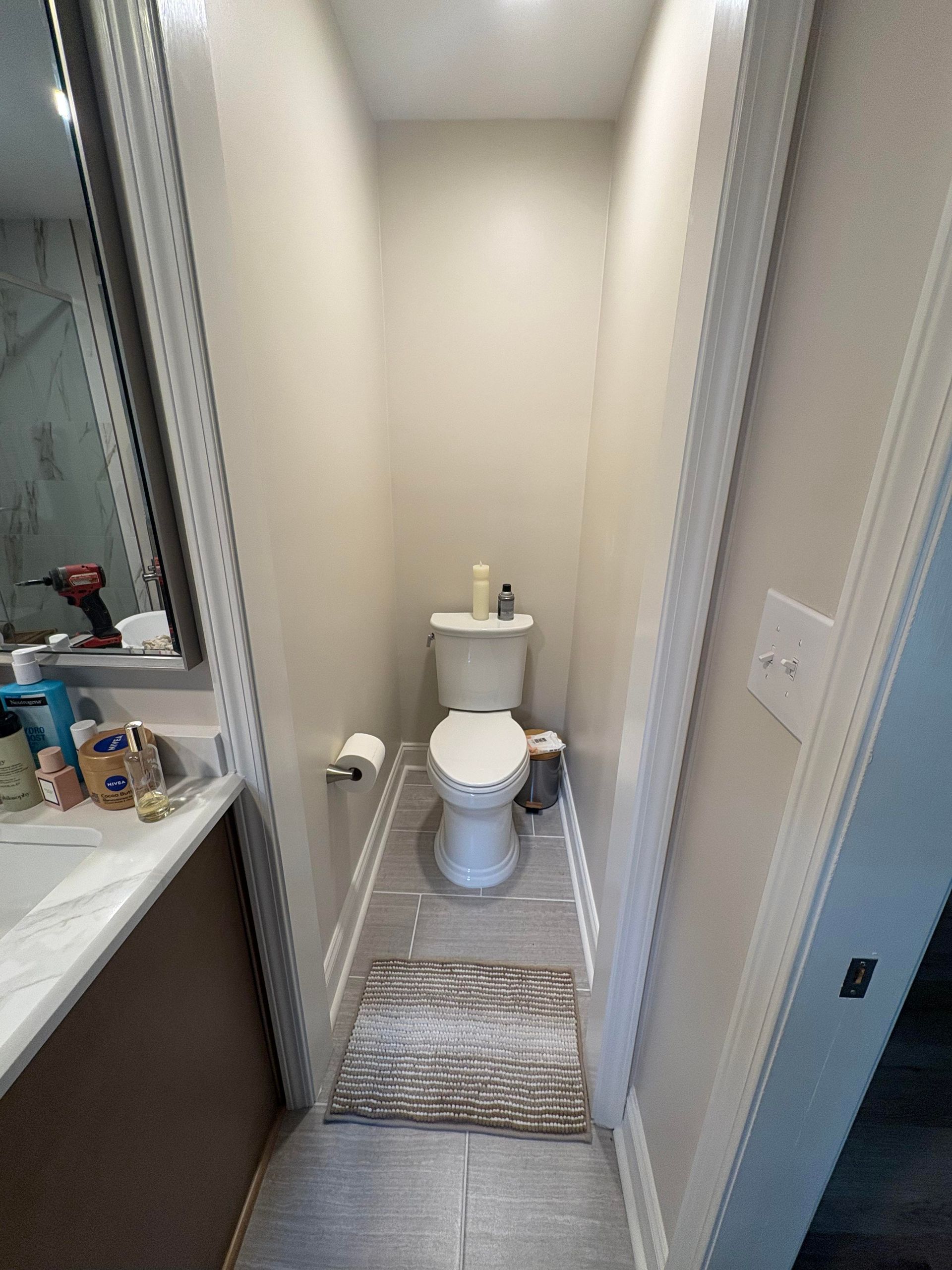 Narrow bathroom with toilet, light gray tile floor, beige walls, and a small rug.