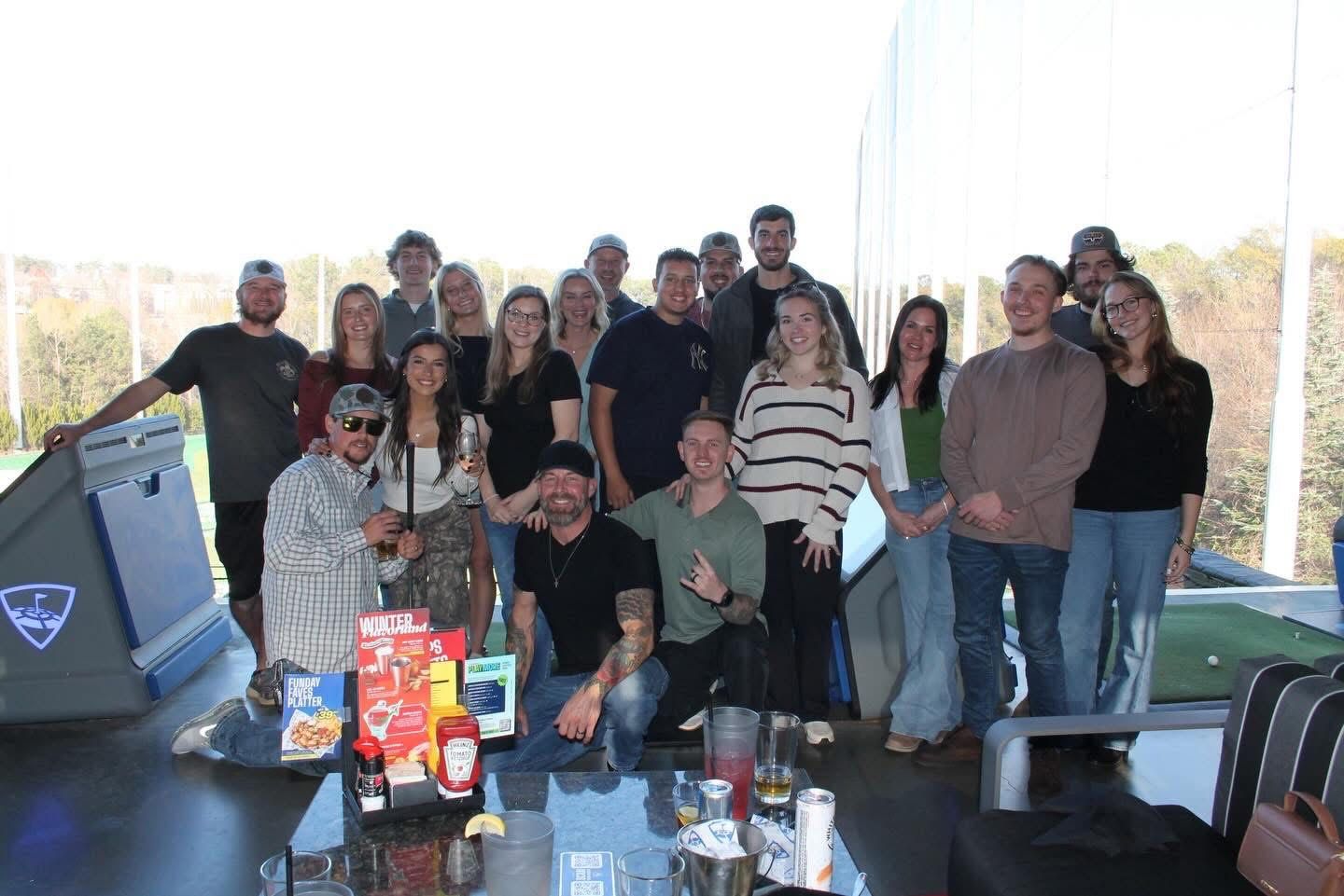 Group of people at a Topgolf, posing near a table with drinks and food. Blue skies, sunny day.
