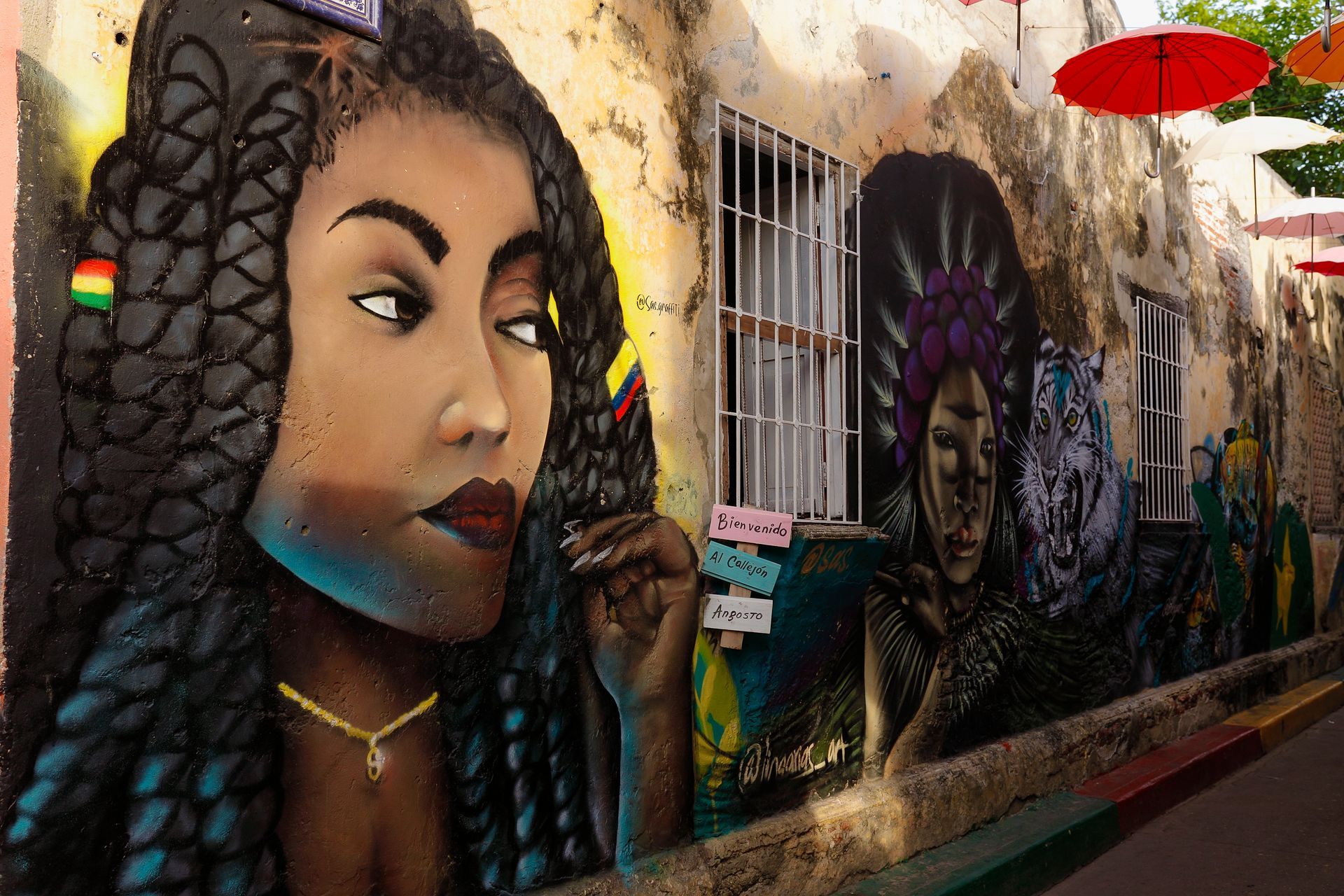 Mural de arte callejero que representa a una mujer con el pelo trenzado, sobre una pared texturizada con detalles coloridos y sombrillas colgantes.