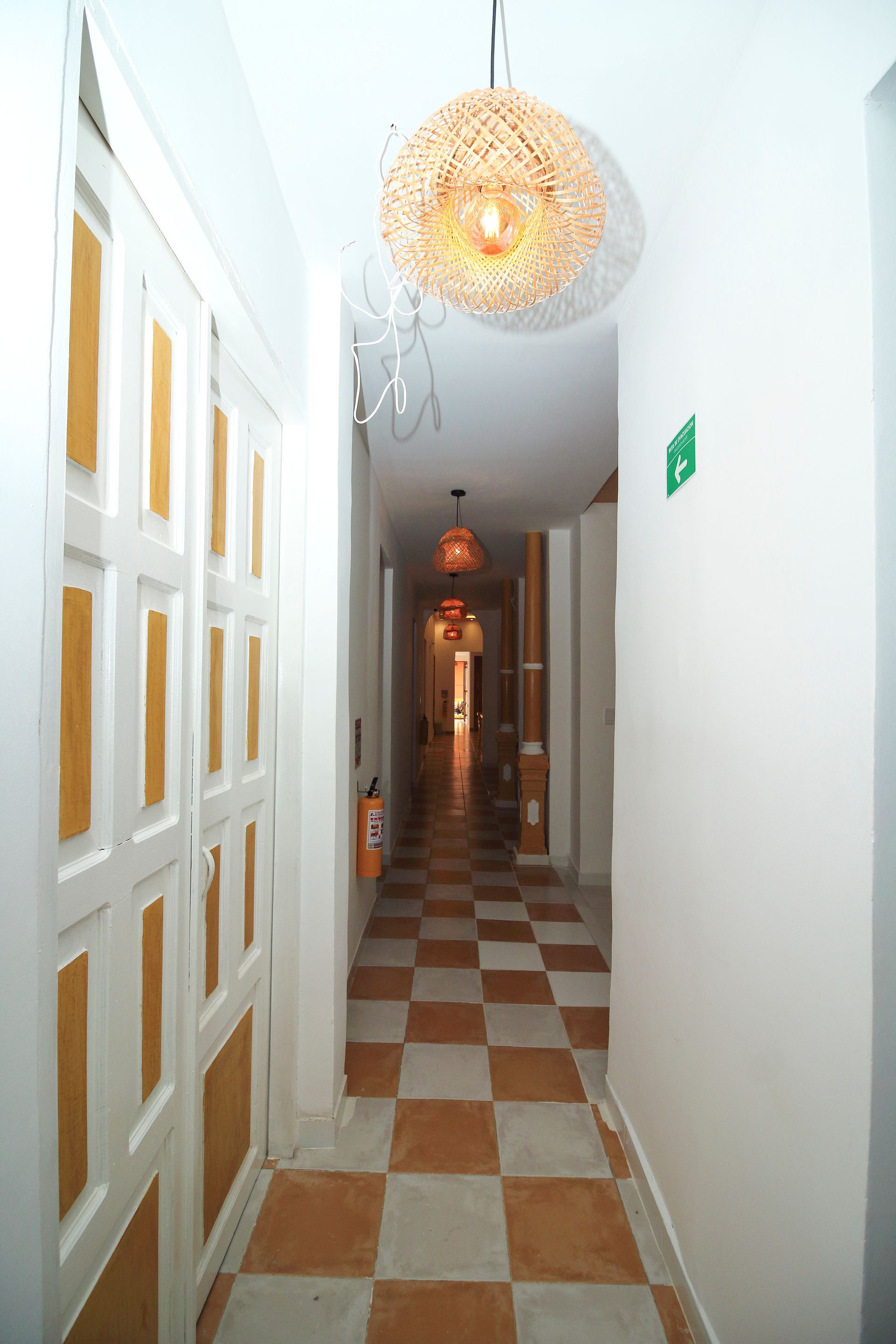 Largo pasillo con suelo ajedrezado, paredes blancas y lámparas colgantes esféricas.