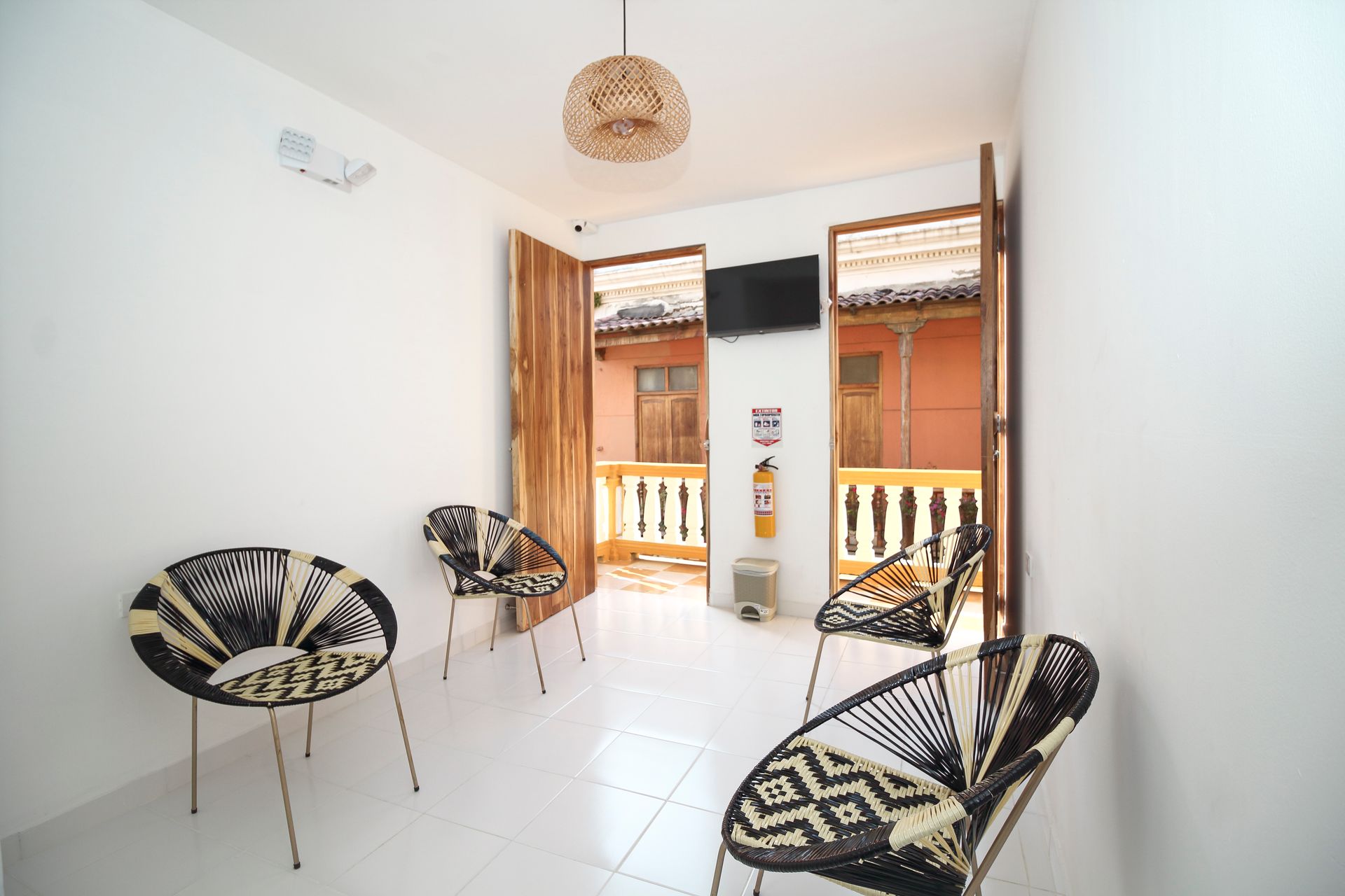 Una pequeña habitación con sillas de mimbre, puertas abiertas a un balcón, un televisor y una lámpara decorativa.