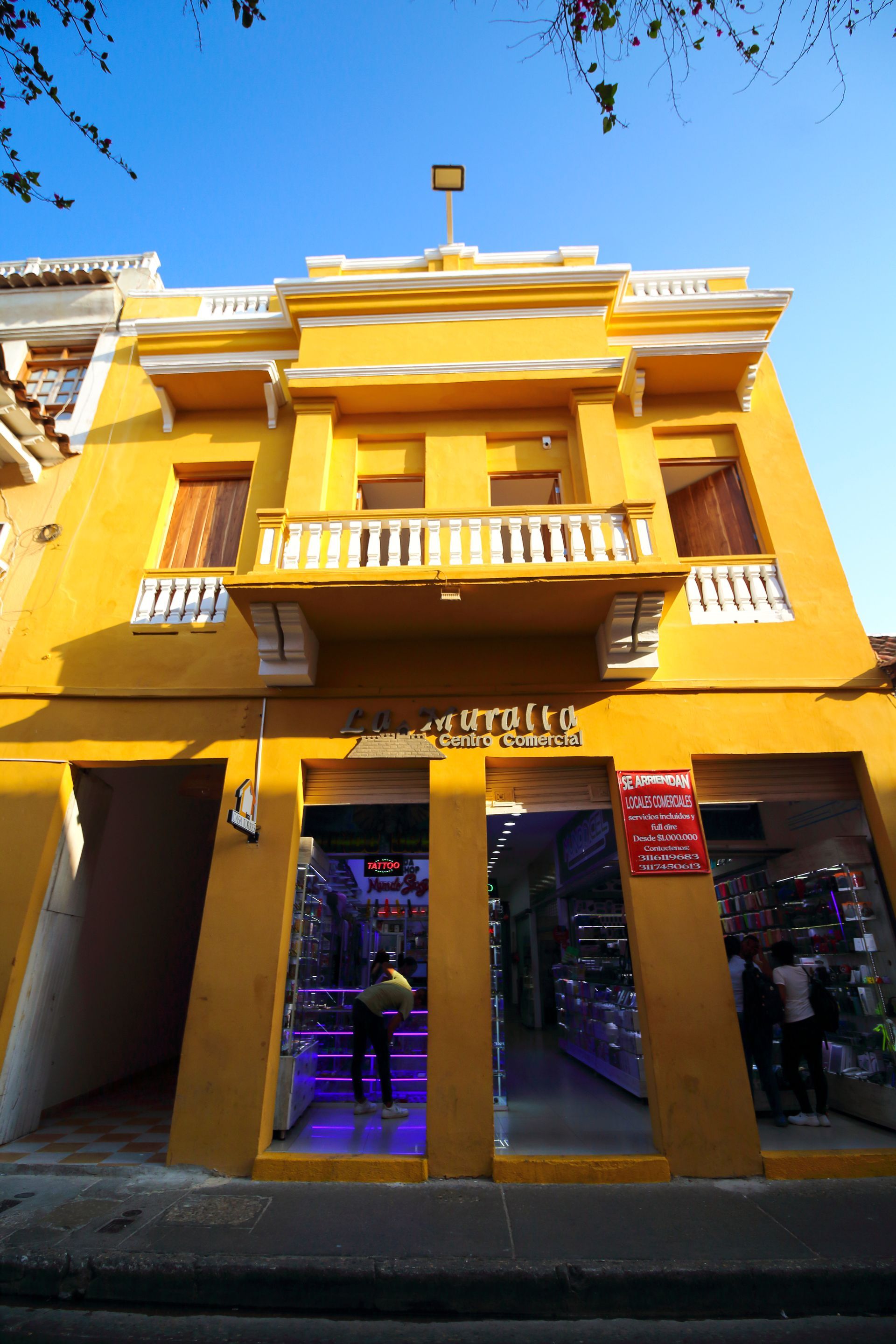 Edificio amarillo con tiendas debajo del balcón, escaparates en la parte inferior, cielo azul arriba.