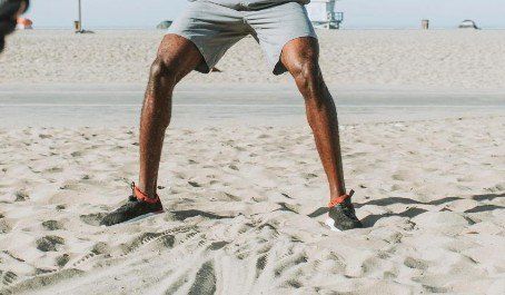 A person shown from shins down wearing black shoes on a beach. A person shown from shins down stands on a beach wearing black shoes.
