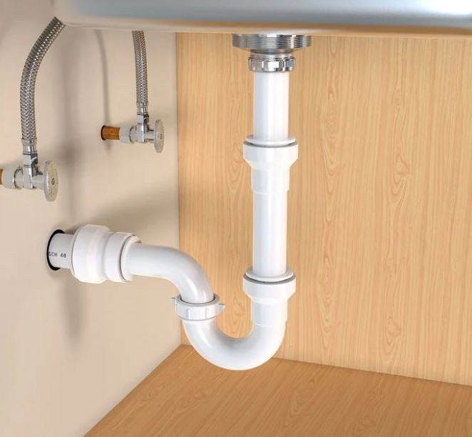 Plumbing under a sink: white pipes, including a P-trap, connected to a drain & water supply valves against a wooden cabinet.