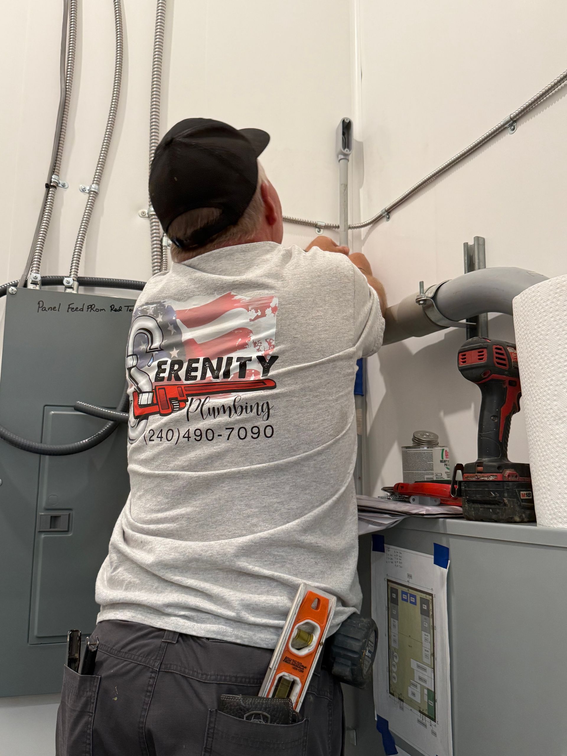 Man in gray shirt installing electrical conduit on a wall; the shirt has a company logo and phone number.