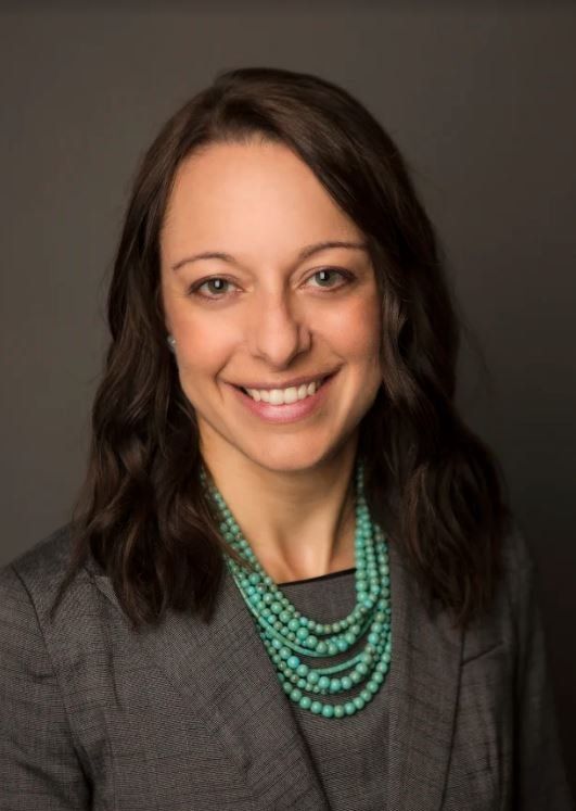 A woman wearing a grey jacket and a green necklace smiles for the camera