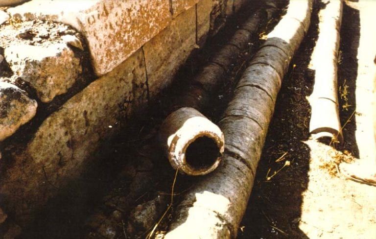A bunch of pipes are laying in the dirt near a brick wall