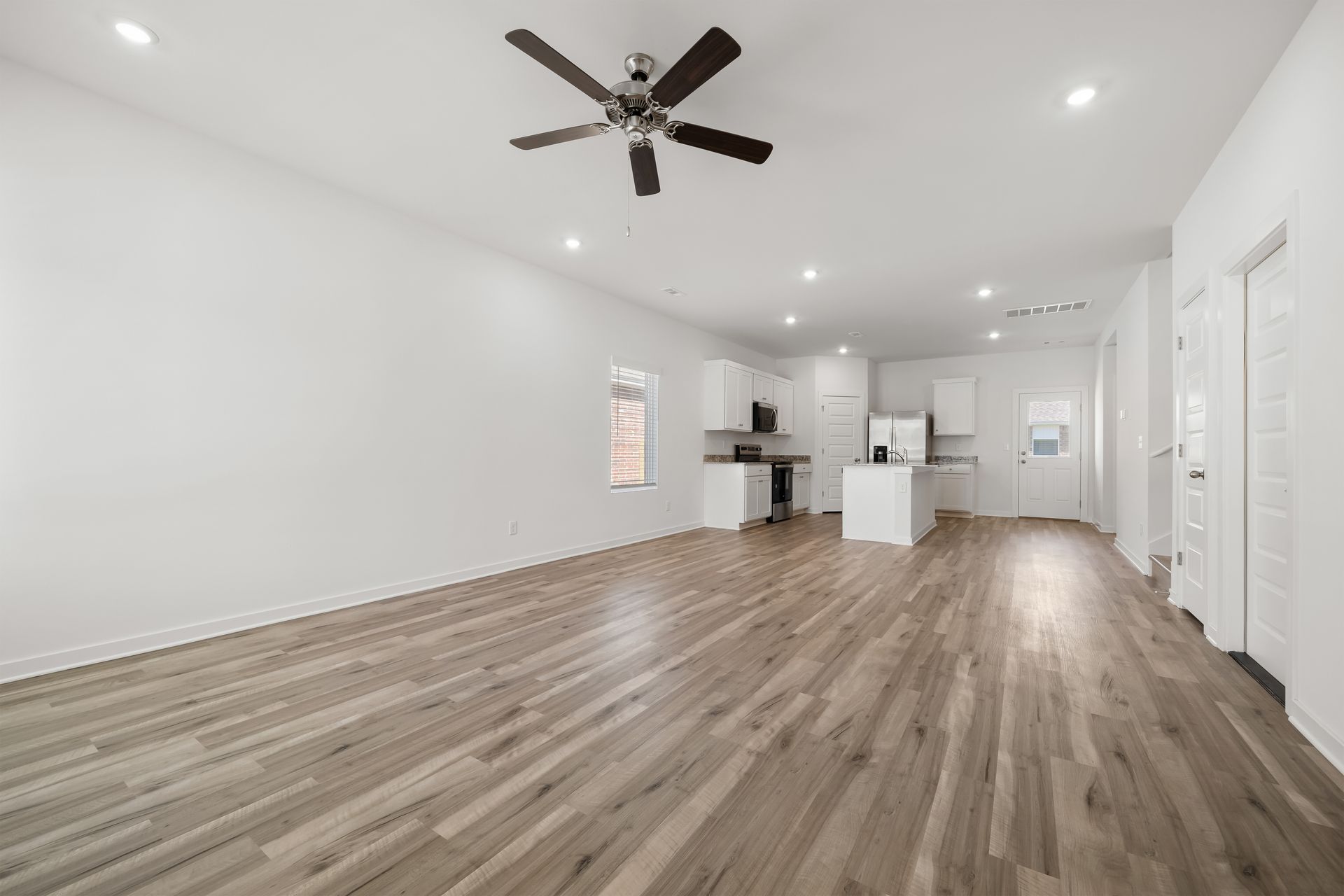 Spacious, empty living area with wood floors, white walls, and a kitchen in the background. Ceiling fan and recessed lights.
