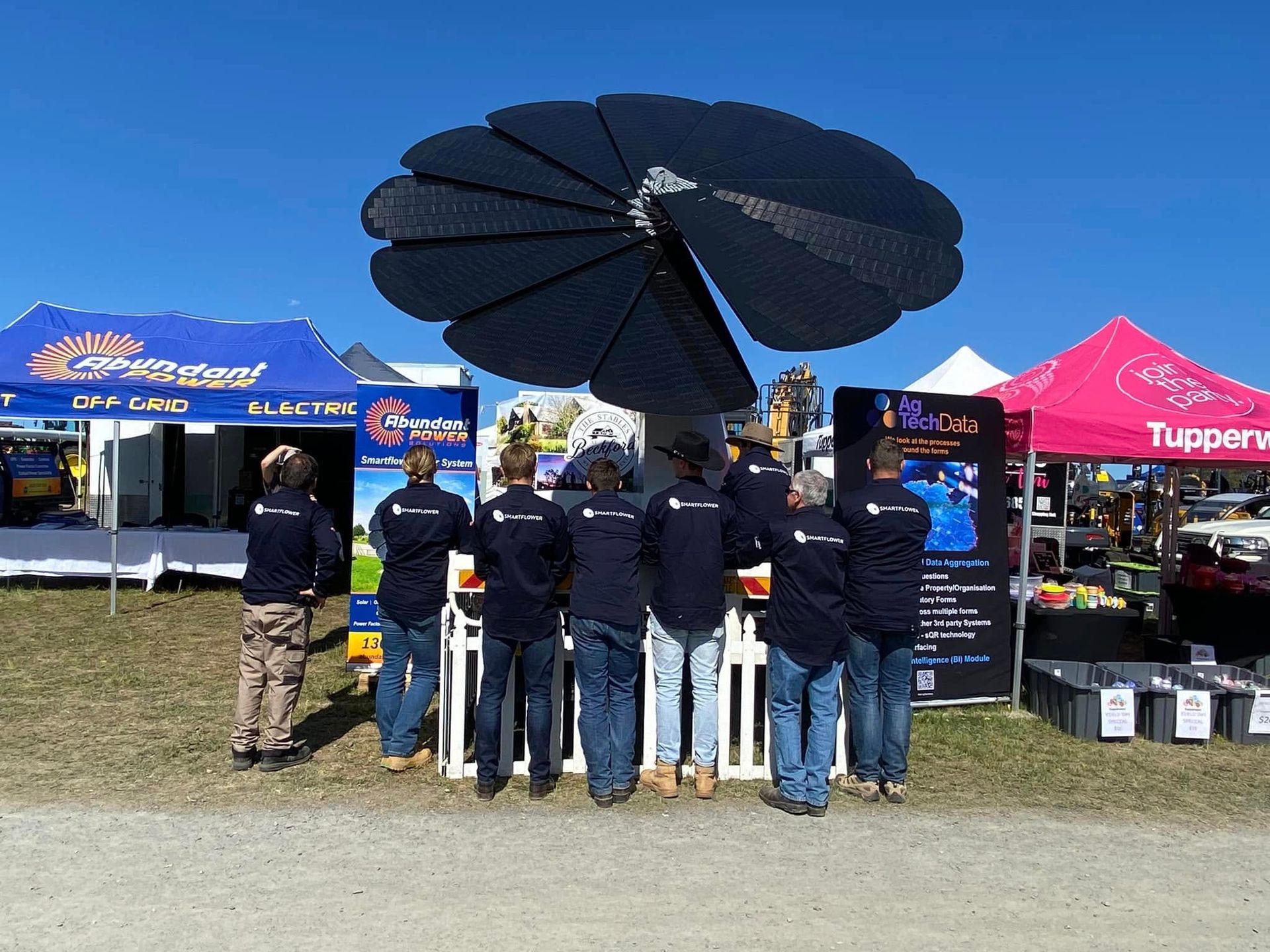Group of people under a large black fan display at an outdoor event booth on a sunny day