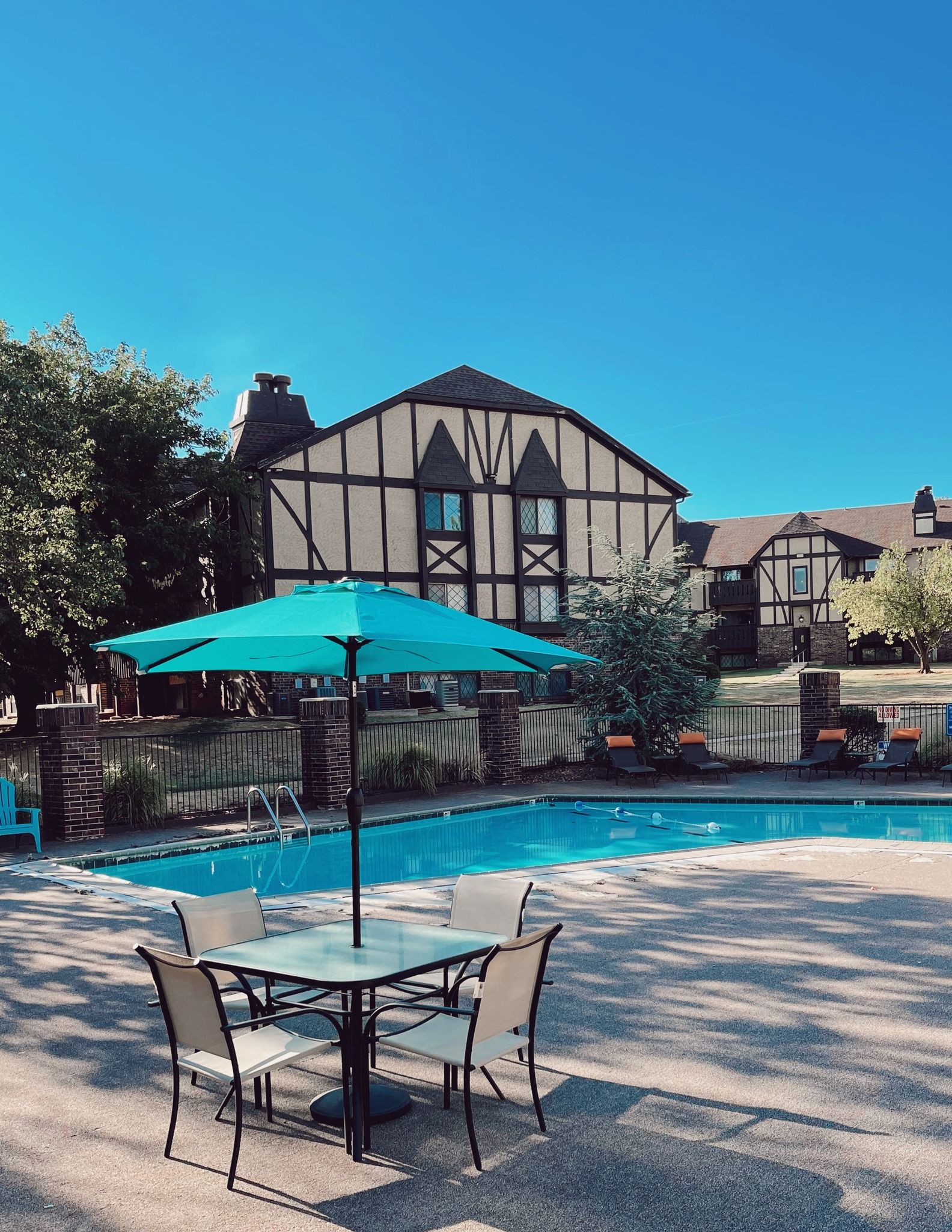 Poolside scene with table, chairs, teal umbrella, and Tudor-style apartment building.
