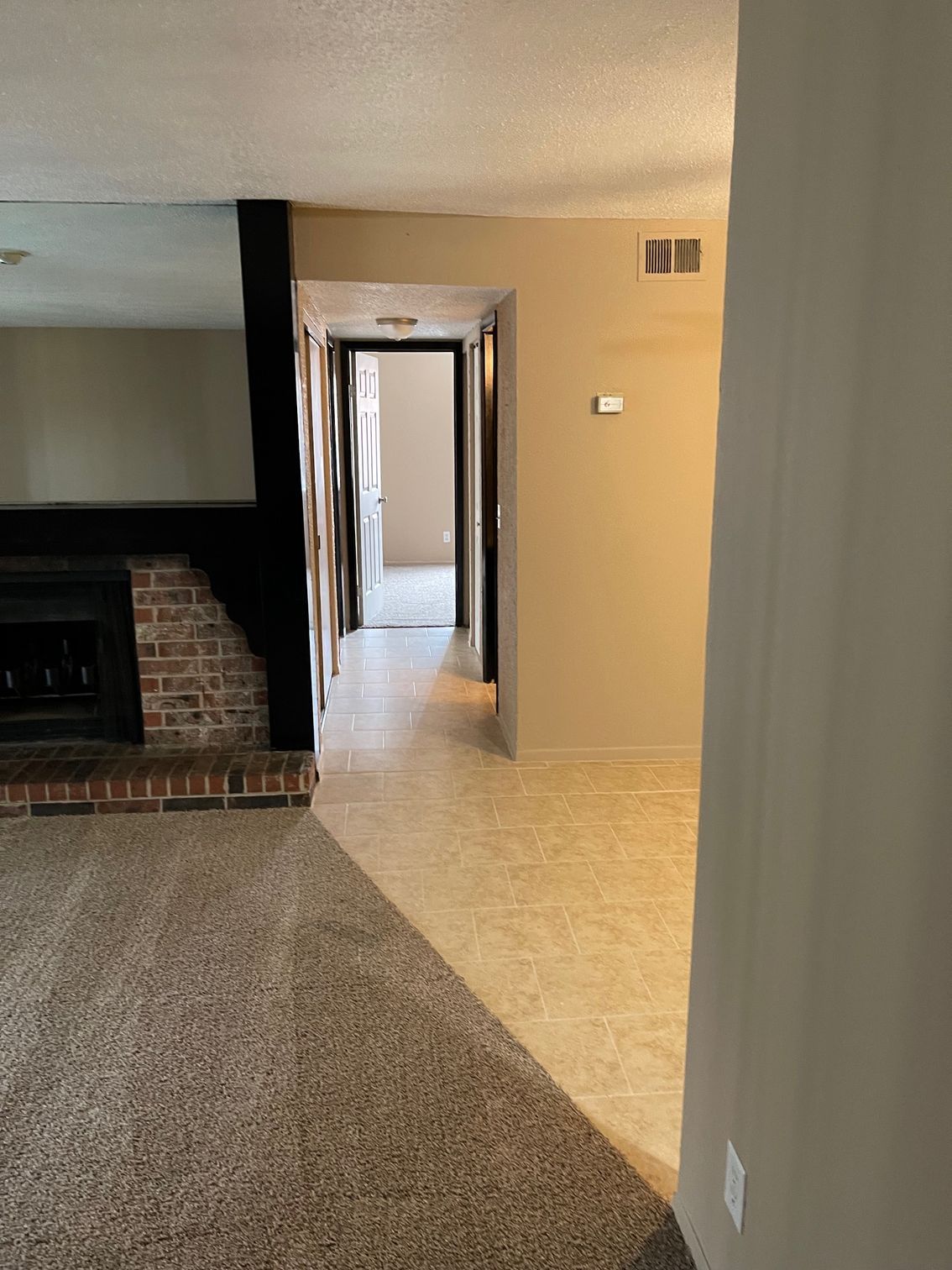 Hallway with beige walls, tile floor, leading to a doorway. Carpeted area with fireplace on left.