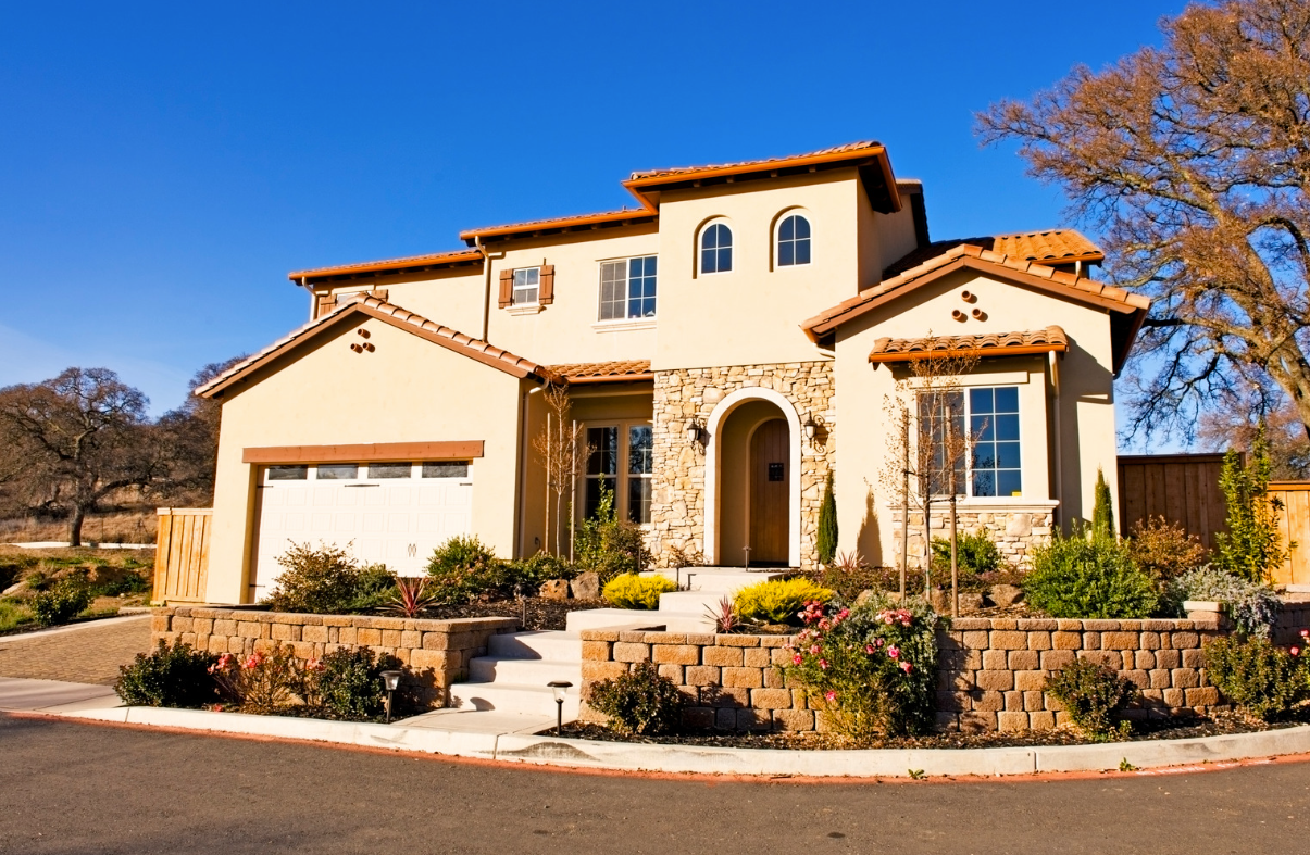 Two-story stucco house with terracotta roof and stone accents, landscaping, and clear blue sky.