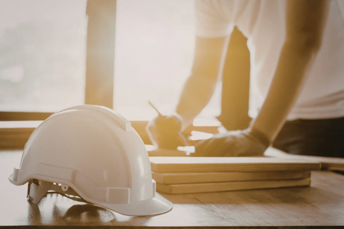 White hard hat on a wooden table, with a person in the background working with wood.