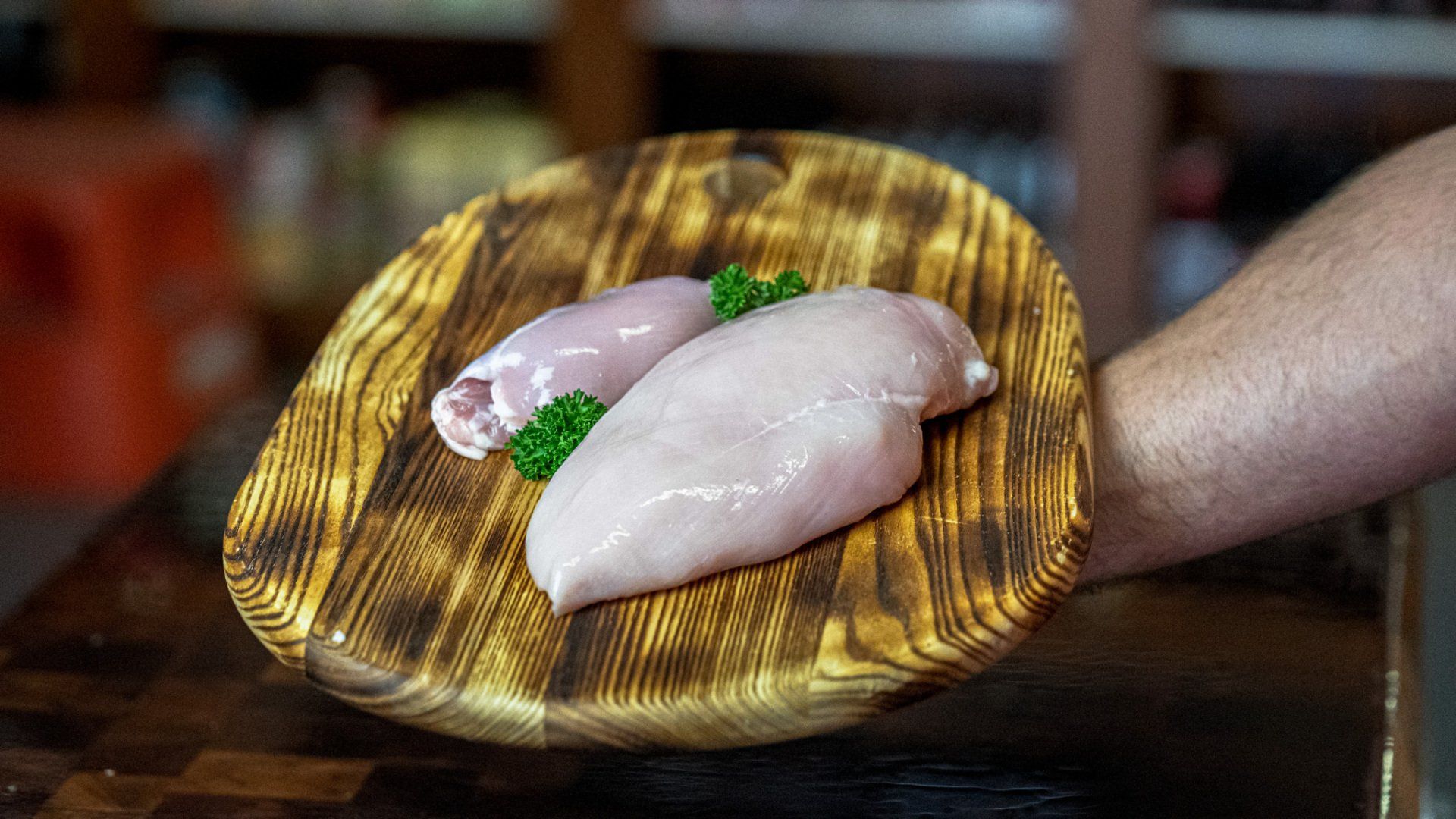 A person is holding a wooden cutting board with two chicken breasts on it.