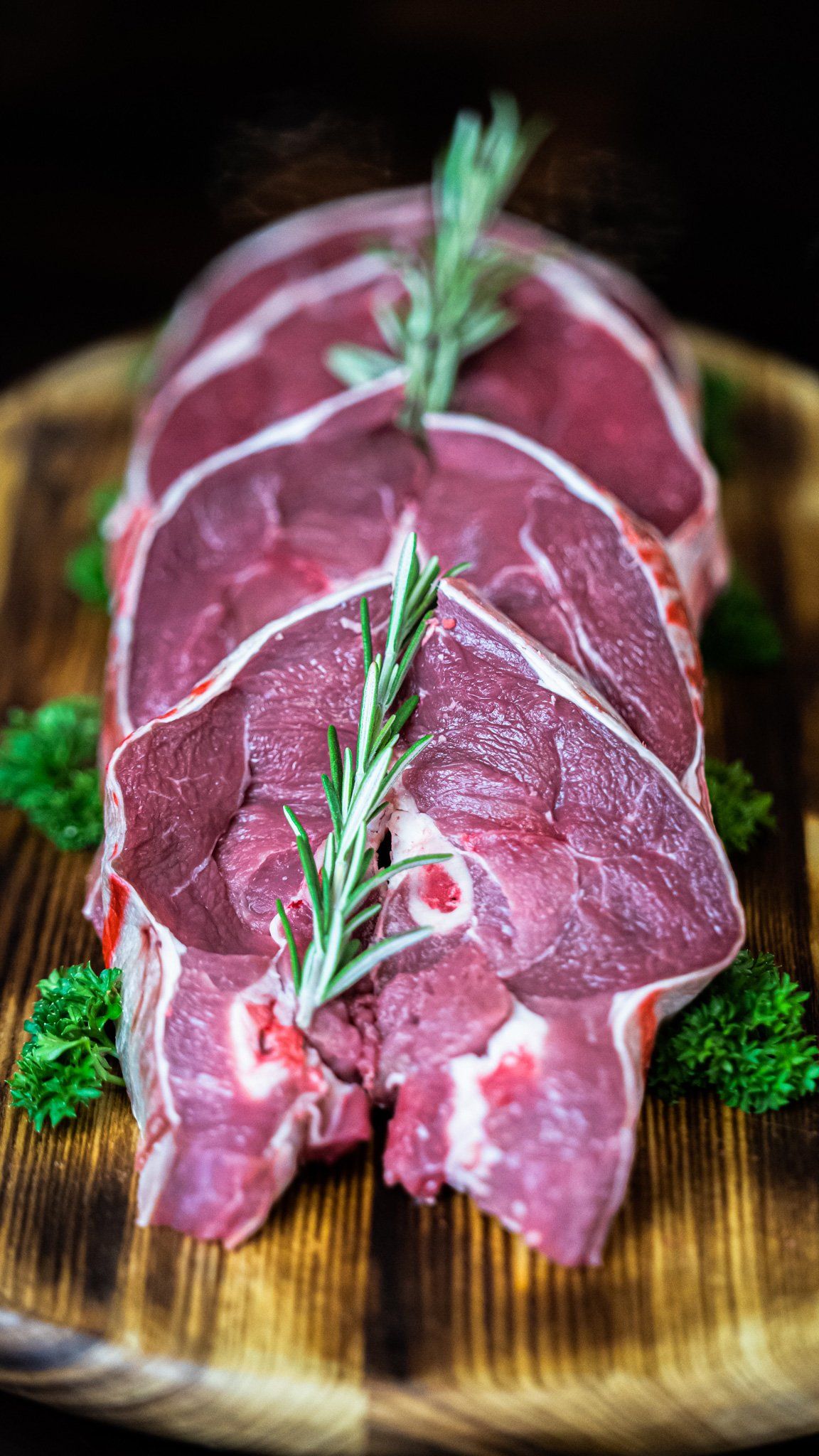 Raw lamb chops on a wooden cutting board with rosemary and parsley.