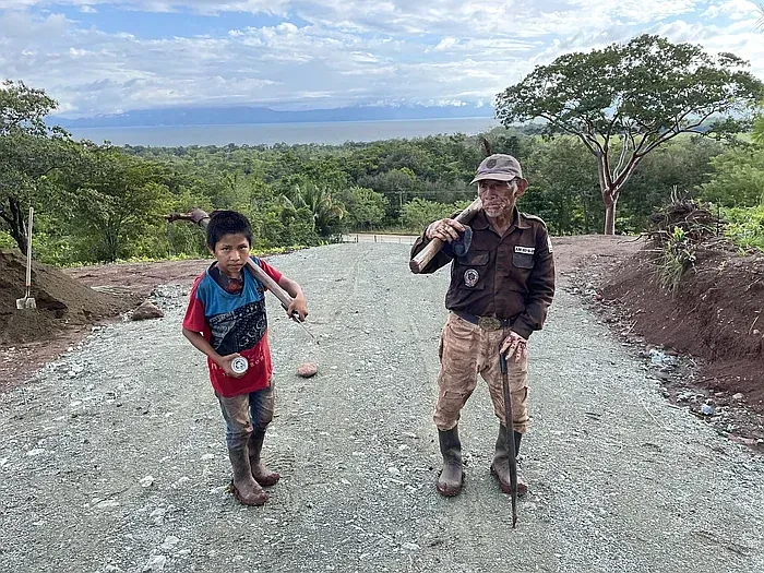 Boy and man stand on a gravel road, holding tools. Lush green landscape and water in the background.