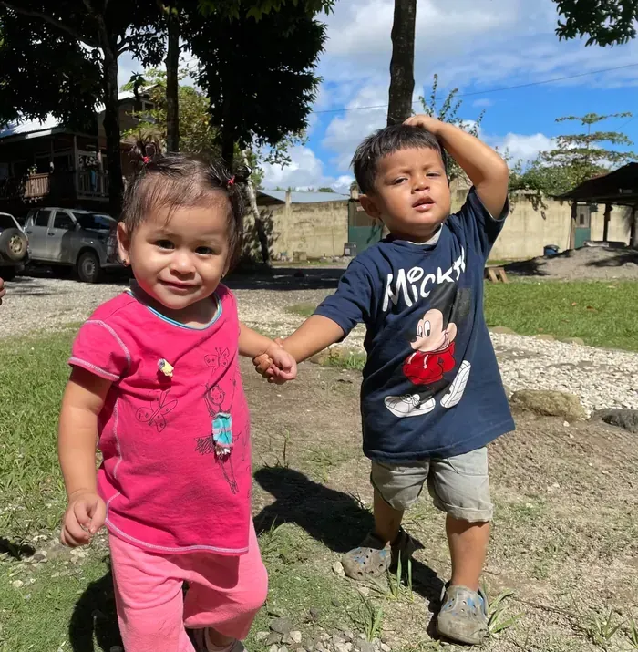 Two young children, a girl in pink and boy in a Mickey Mouse shirt, holding hands, smiling outside on a sunny day.