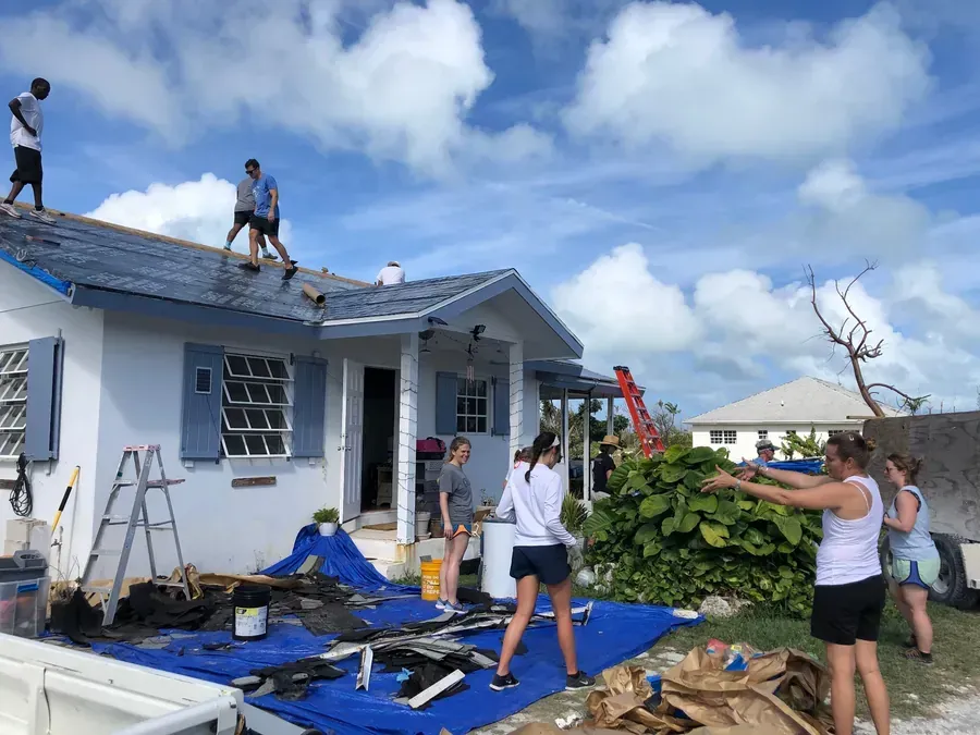 People repairing a roof and cleaning up debris near a small house under a cloudy sky.