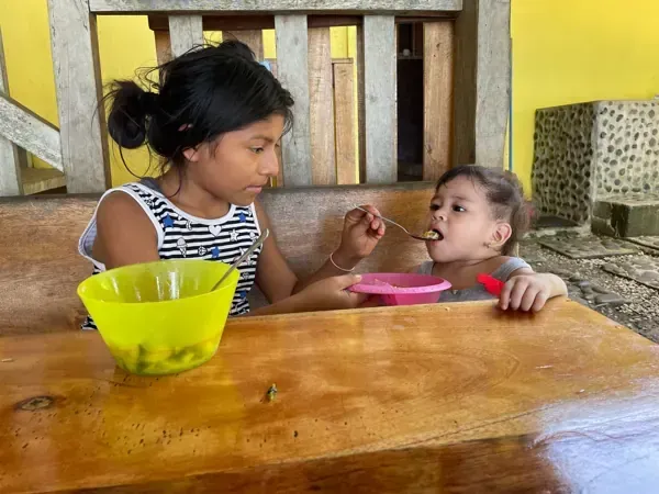 A girl feeds another girl at a wooden table. Yellow bowl and pink plate.