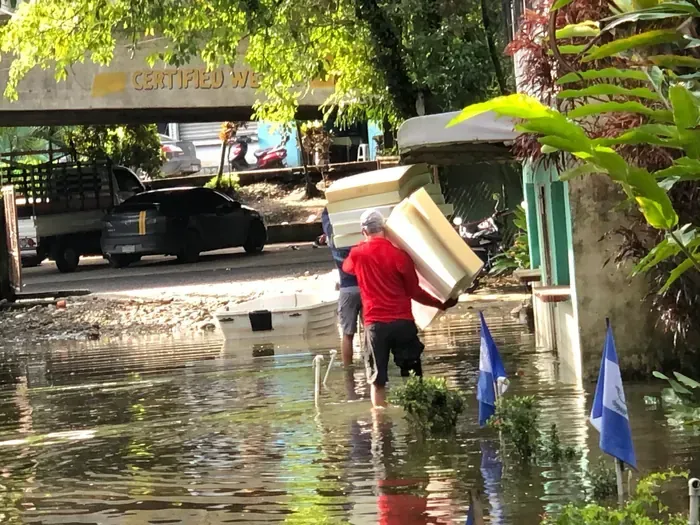 People wading through floodwater, carrying a large object, near a building with flags.