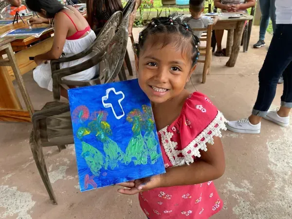 Girl smiles, holding a blue painting with green figures and a white cross. Outdoors.