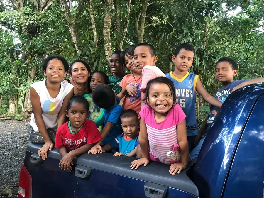 Group of children and adults smiling in the back of a blue truck, surrounded by trees.