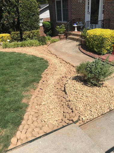 Curved concrete walkway bordered by tan rock landscaping, green grass, shrubs, and a house entrance.