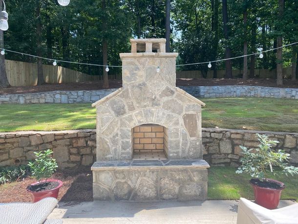 Stone outdoor fireplace in a landscaped yard, with potted plants and a stone retaining wall behind it