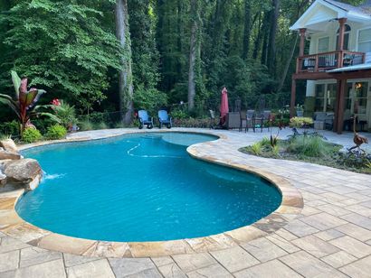 Curved backyard pool with blue water, stone patio, lounge chairs, and a house beside dense trees