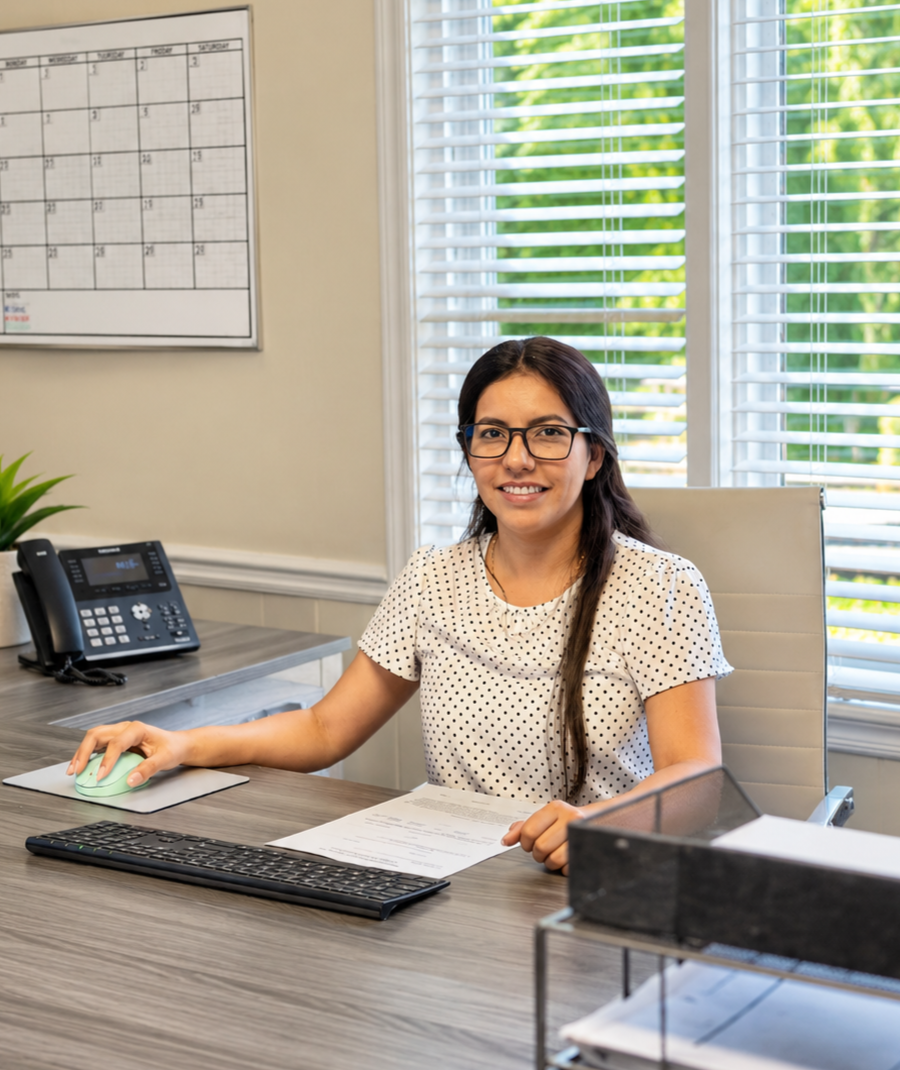 Woman seated at an office desk with a computer, phone, and blinds-covered windows behind her.