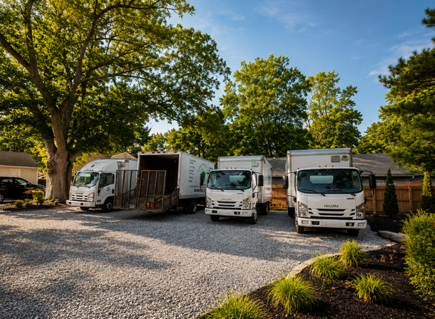 Several white trucks parked on gravel under trees, with a cloudy sky overhead.