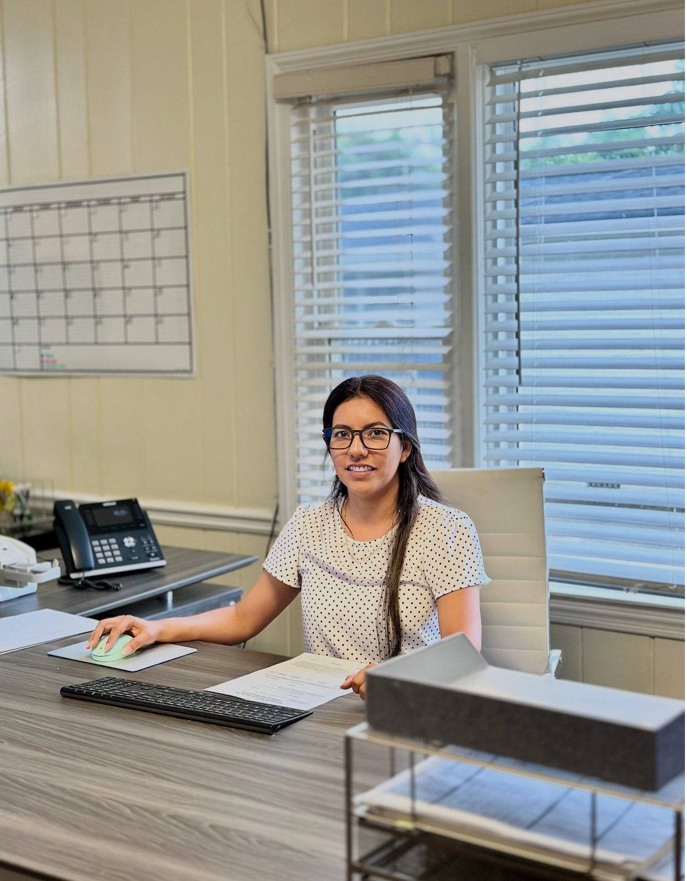 Woman seated at an office desk with a computer, phone, and blinds-covered windows behind her.