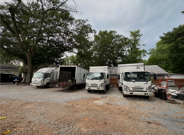 Several white trucks parked on gravel under trees, with a cloudy sky overhead.
