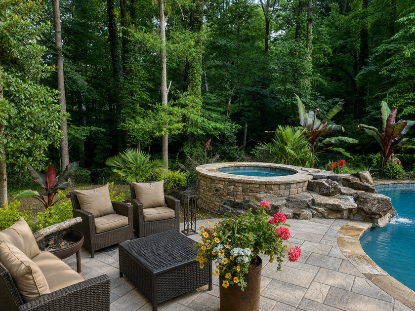Patio seating beside a stone hot tub and pool in a wooded backyard