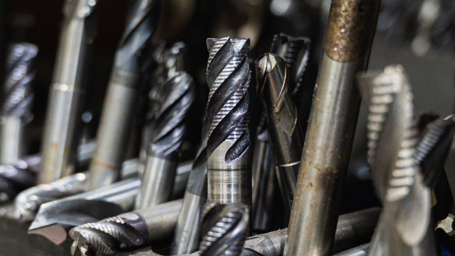 Close-up of a collection of various metal drill bits, mostly silver, some with dark fluting, in a workshop.