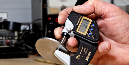 A hand using a digital micrometer to measure the thickness of a small, circular metal object.