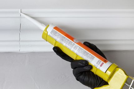 A person wearing black gloves, applying caulk to a crack in white molding with a yellow caulking gun.