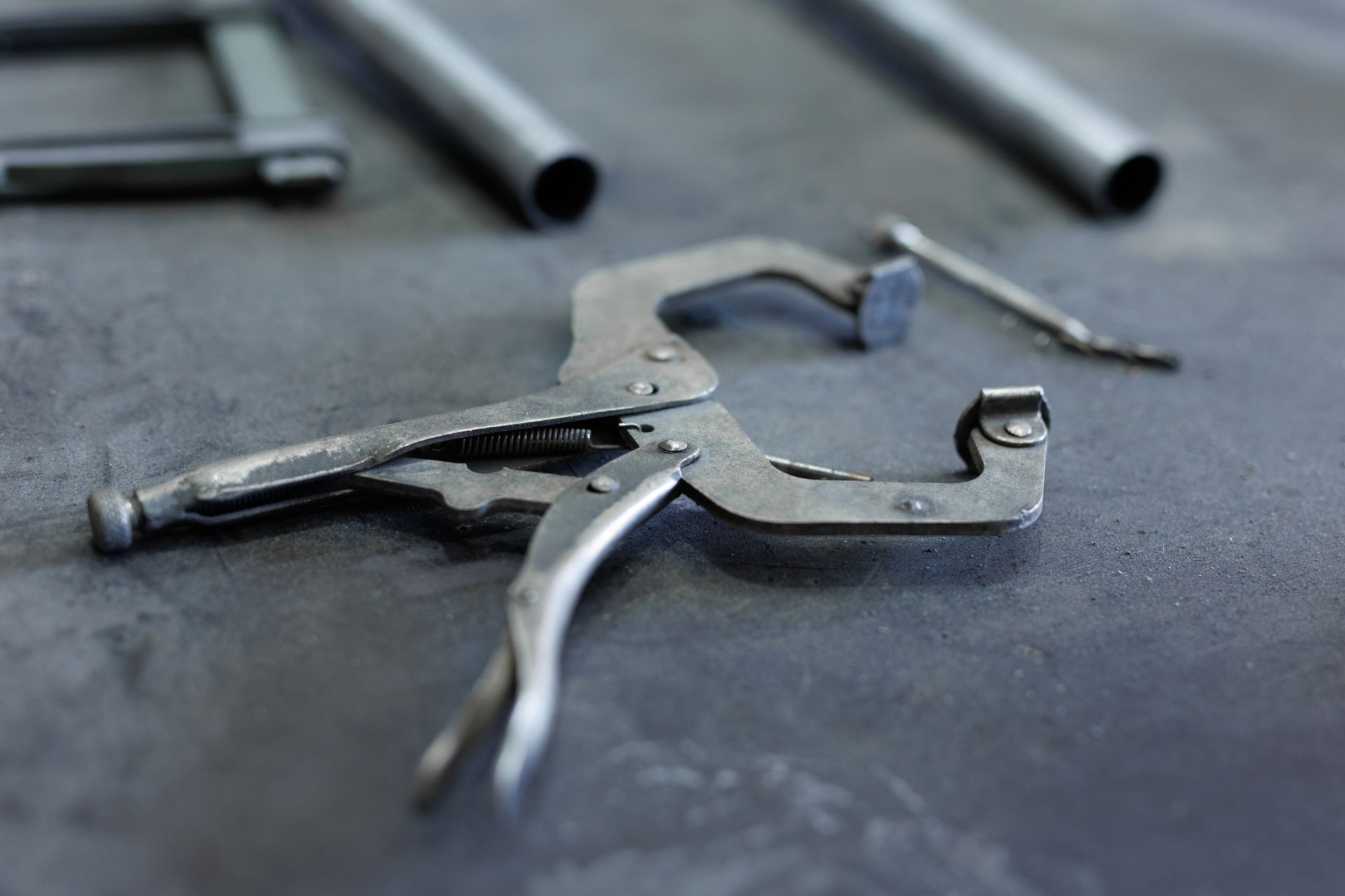 A locking clamp with two metal tubes and a smaller clamp in the background on a dark surface.