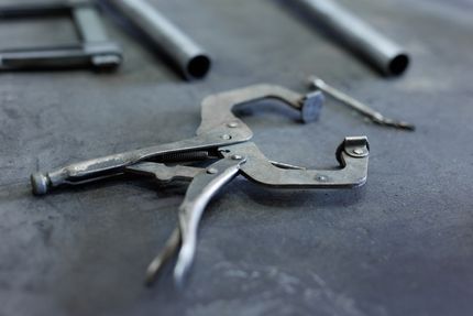 Metal locking pliers next to metal tubes, all on a dark surface.
