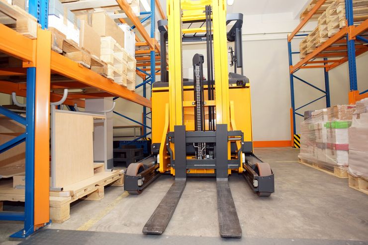 Yellow forklift in a warehouse aisle, facing forward with forks extended, ready to lift pallets.