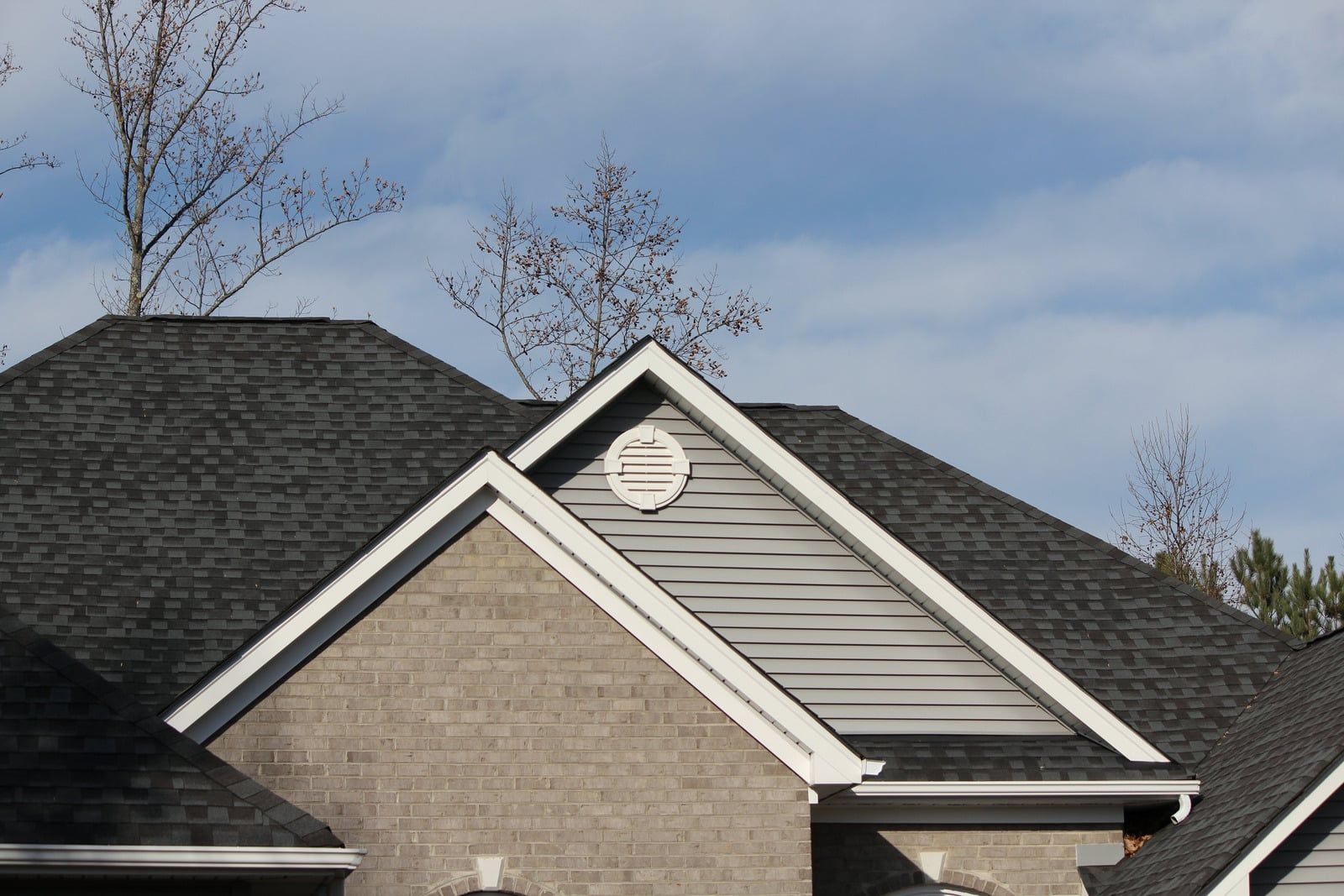 Suburban house roof and gable with beige siding under a cloudy blue sky
