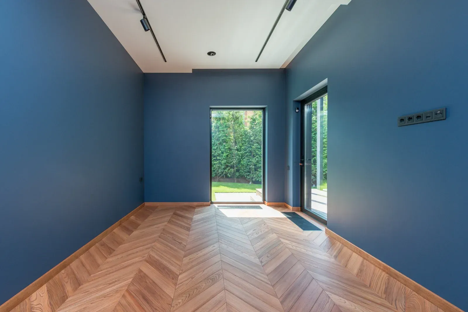 Empty blue room with herringbone wood floor and glass doors opening to a green garden.