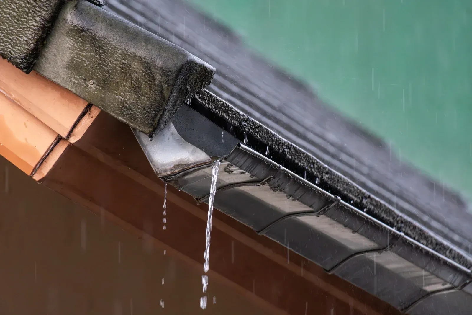 Rainwater dripping from a roof gutter beside orange tiles and a green wall