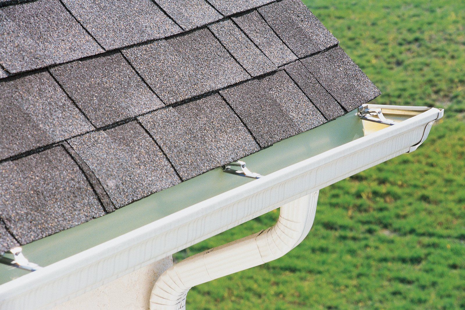 Roof gutter with a green leaf guard beside gray shingle roof and white downspout