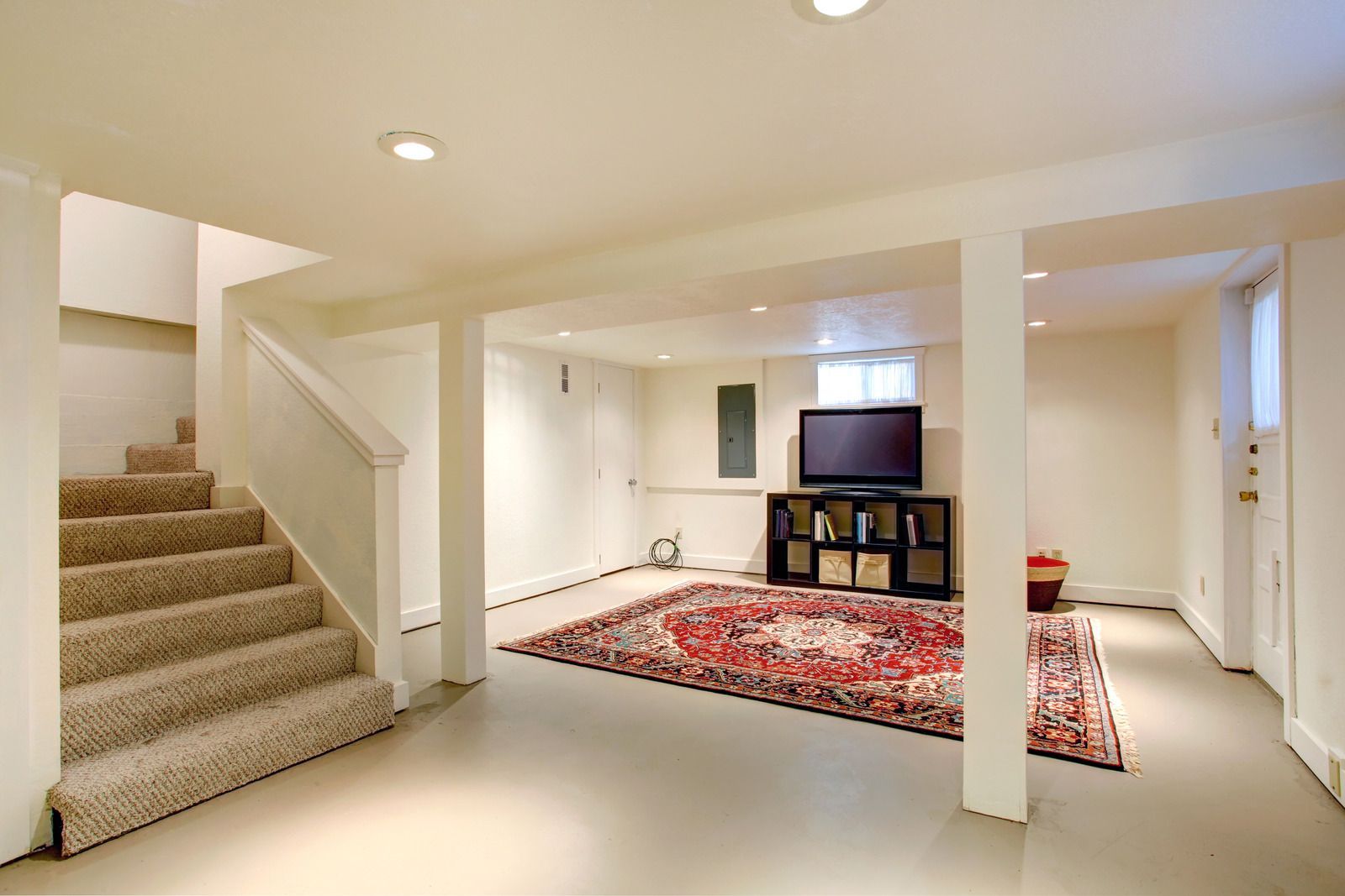 Basement living room with beige carpet stairs, white columns, and a red rug with a TV on a shelf unit