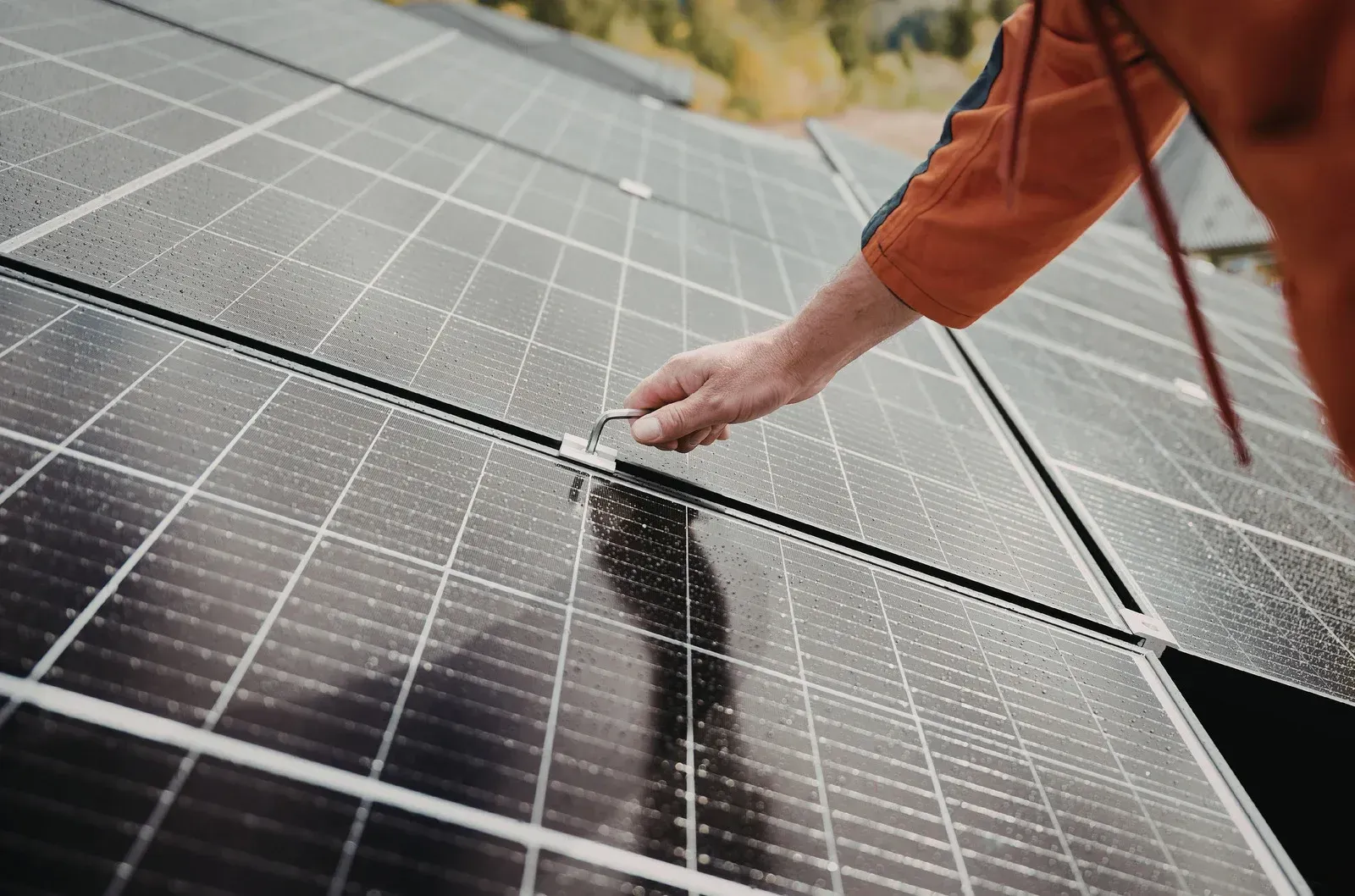 Person cleaning solar panels with a squeegee on a rooftop, wearing an orange jacket.